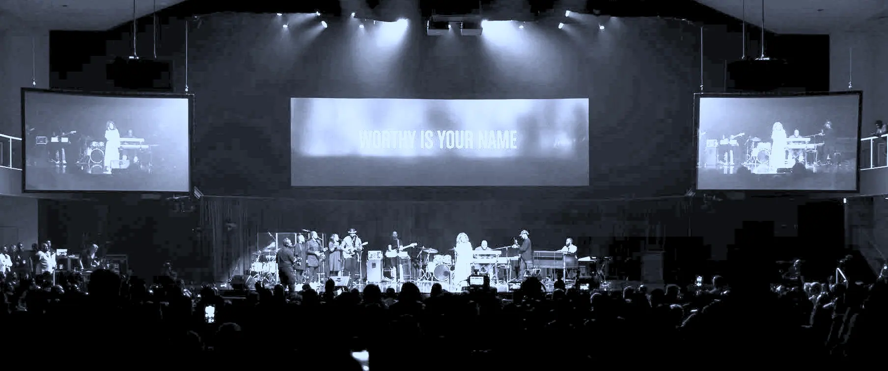 The full hall from the back, three screens above the band and the song title across the centre one.