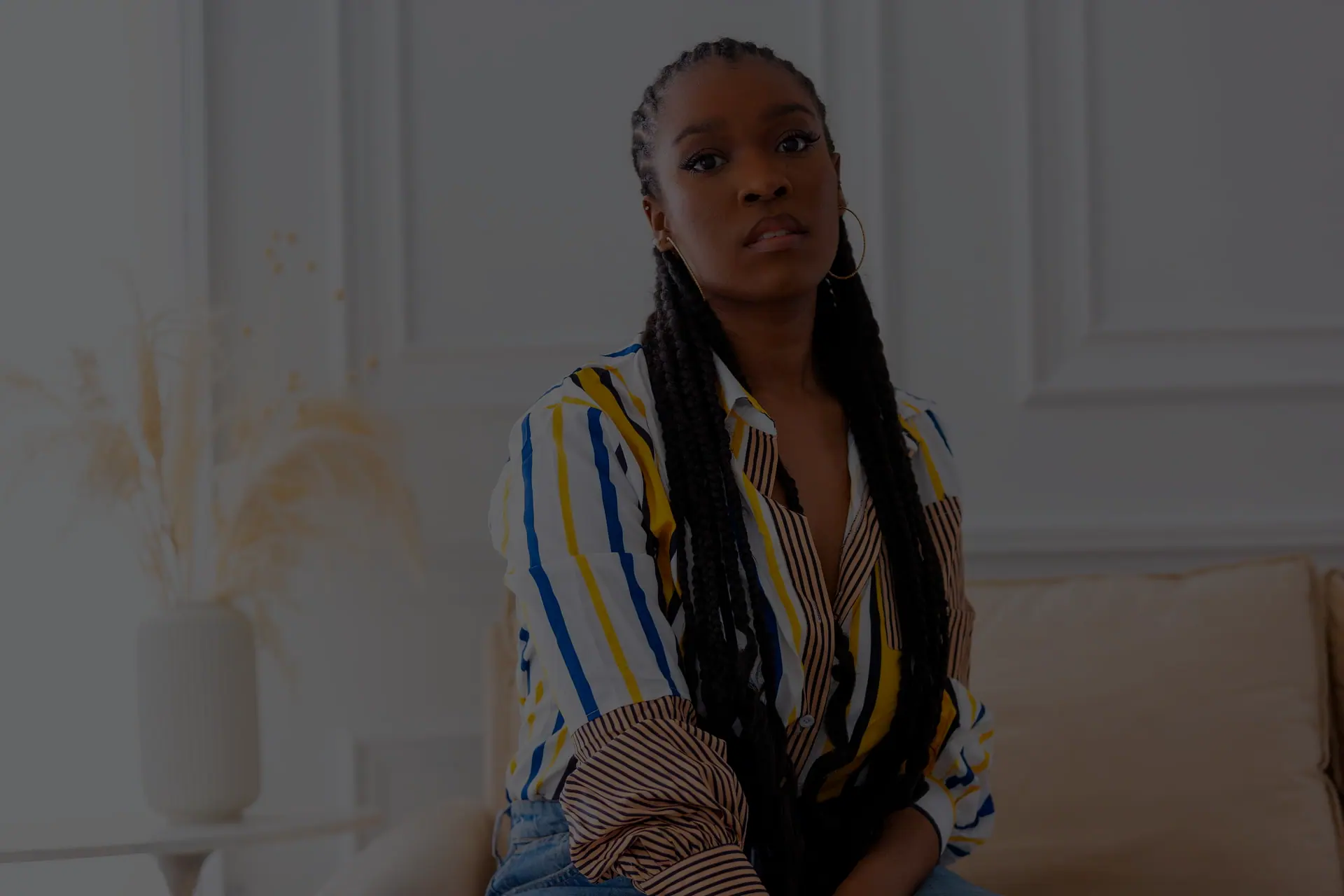 A model on a sofa in a striped shirt, hair in long braids, a bright room behind her.