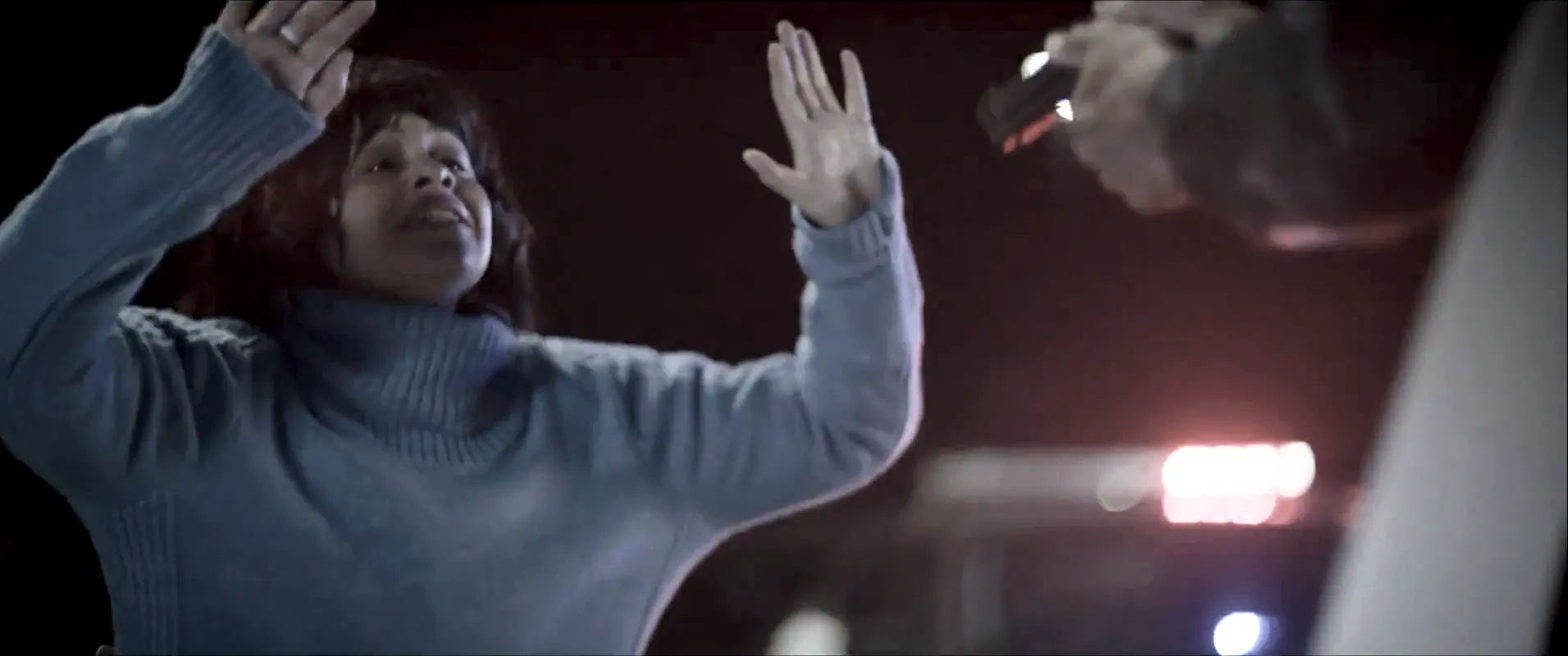 A woman with both hands raised, police lights washing the frame.